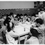 Infants treballant i jugant al voltant d’una taula en una aula de les escoles Garbí Pere Vergés, fotografia històrica en blanc i negre.