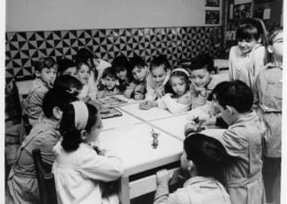 Infants treballant i jugant al voltant d’una taula en una aula de les escoles Garbí Pere Vergés, fotografia històrica en blanc i negre.