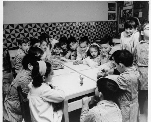 Infants treballant i jugant al voltant d’una taula en una aula de les escoles Garbí Pere Vergés, fotografia històrica en blanc i negre.