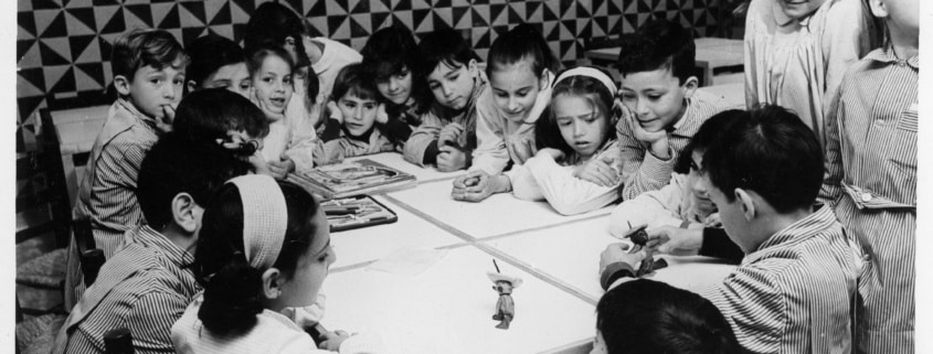 Infants treballant i jugant al voltant d’una taula en una aula de les escoles Garbí Pere Vergés, fotografia històrica en blanc i negre.