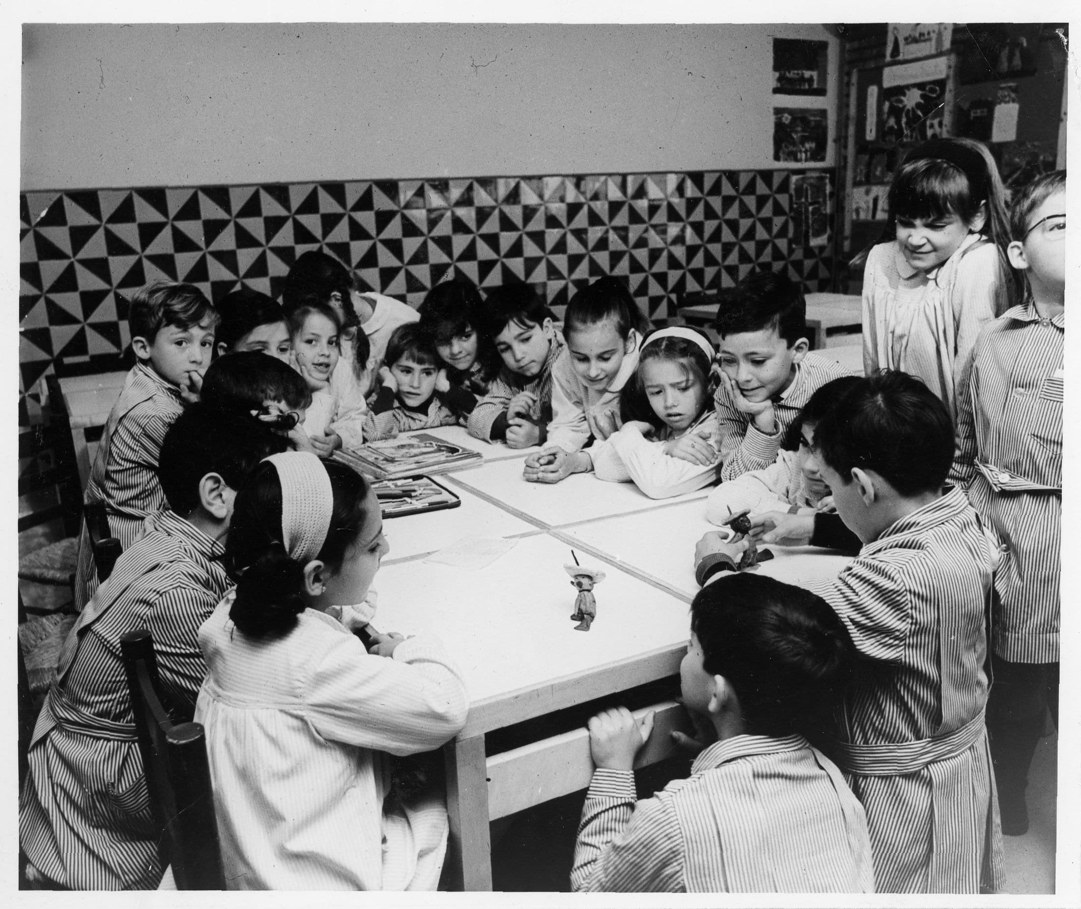 Infants treballant i jugant al voltant d’una taula en una aula de les escoles Garbí Pere Vergés, fotografia històrica en blanc i negre.